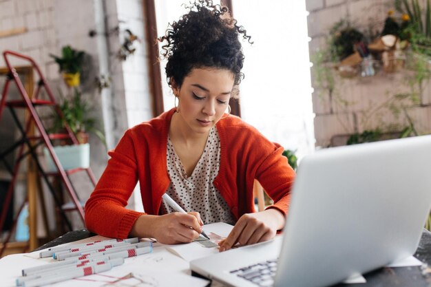 Jovem mulher bonita com cabelo escuro encaracolado, sentado à mesa com laptop sonhadoramente desenhando ilustrações de moda, passando tempo na oficina moderna e aconchegante com grandes janelas