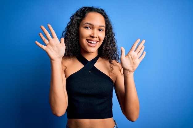 Foto grátis jovem mulher bonita com cabelos cacheados vestindo camiseta casual em pé sobre fundo azul mostrando e apontando para cima com os dedos número dez enquanto sorria confiante e feliz