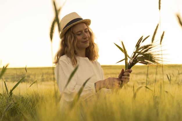 Foto grátis jovem mulher bonita com cabelos longos loiros em um vestido branco com um chapéu de palha coleta flores em um campo de trigo. cabelo voando ao sol, verão. tempo para sonhadores, pôr do sol dourado.