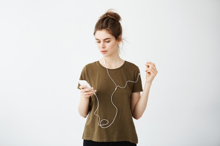 Foto grátis jovem mulher bonita com coque sorrindo olhando para celular ouvindo música em fones de ouvido