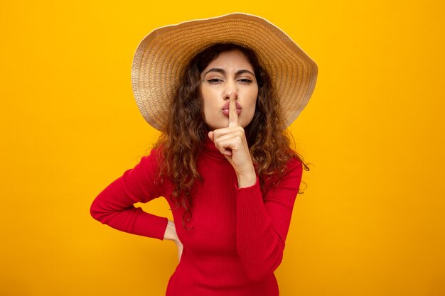 Jovem mulher bonita com gola alta vermelha e chapéu de verão fazendo gesto de silêncio com o dedo nos lábios em pé na laranja