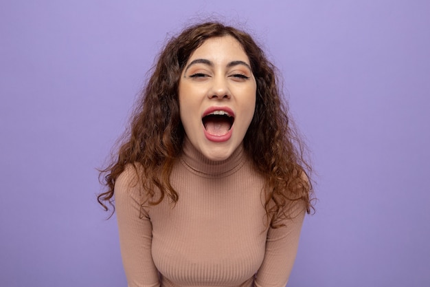 Jovem mulher bonita com gola rulê bege gritando bem alto e animada e feliz