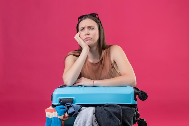 Foto grátis jovem mulher bonita com mala de viagem cheia de roupas, olhando de lado com o rosto carrancudo descontente e cansado pensando sobre parede rosa