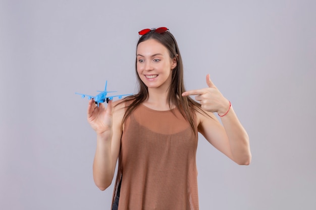 Jovem mulher bonita com óculos de sol vermelhos na cabeça segurando um avião de brinquedo apontando com o dedo para ele, positivo e feliz sorrindo em pé sobre um fundo branco