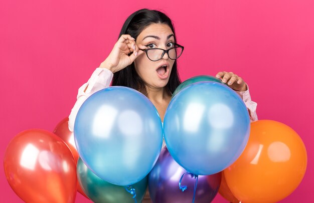 Foto grátis jovem mulher bonita com óculos, olhando para a câmera, em pé atrás de balões isolados na parede rosa