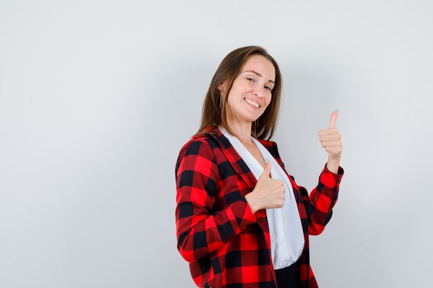 Jovem mulher bonita com roupa casual, mostrando os polegares e olhando bem-aventurada, vista frontal.