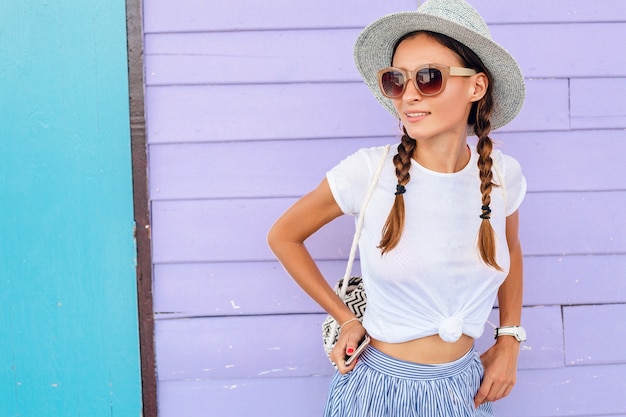 Foto grátis jovem mulher bonita com roupa de moda verão estilo casual, posando contra uma parede colorida, viajando, chapéu, óculos de sol, sorrindo, feliz