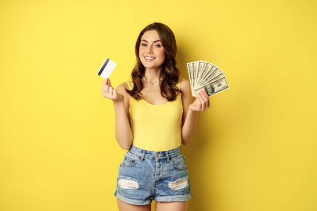 Jovem mulher bonita com roupa de verão, segurando o cartão de crédito e dinheiro, dinheiro nas mãos, de pé contra um fundo amarelo.