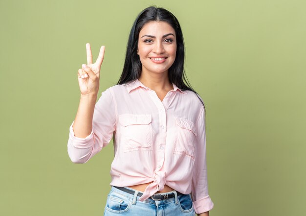 Jovem mulher bonita com roupas casuais, olhando para a frente, sorrindo alegremente, mostrando o número dois com os dedos em pé sobre a parede verde