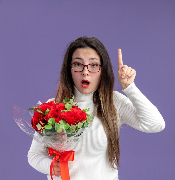 jovem mulher bonita com roupas casuais segurando um buquê de rosas vermelhas olhando para a câmera surpresa, mostrando o conceito do dia dos namorados com o dedo indicador em pé sobre a parede roxa