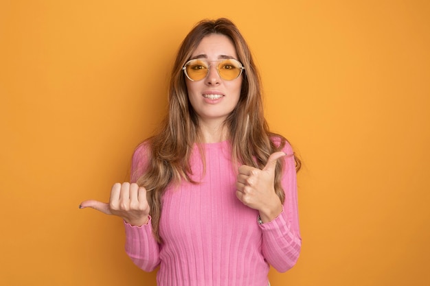 Jovem mulher bonita com top rosa usando óculos, olhando para a câmera, confusa, mostrando os polegares apontando para o lado com o polegar em pé sobre um fundo laranja