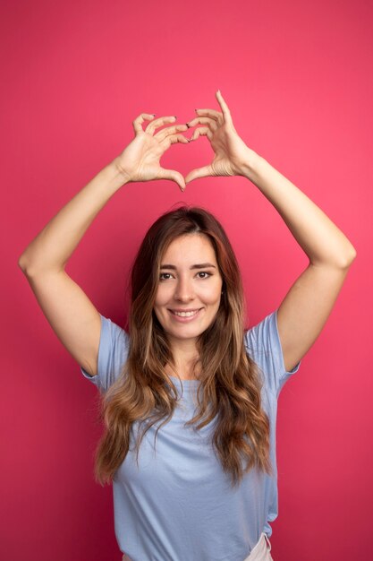 Jovem mulher bonita com uma camiseta azul fazendo um gesto de coração sobre a cabeça e sorrindo alegremente