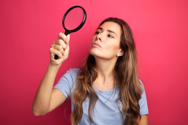 Foto grátis jovem mulher bonita com uma camiseta azul segurando uma lupa e olhando para ela intrigada em pé sobre rosa