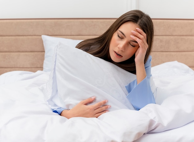 Jovem mulher bonita de pijama azul deitada na cama com travesseiro bocejando, sentindo fadiga matinal no interior do quarto com luz de fundo