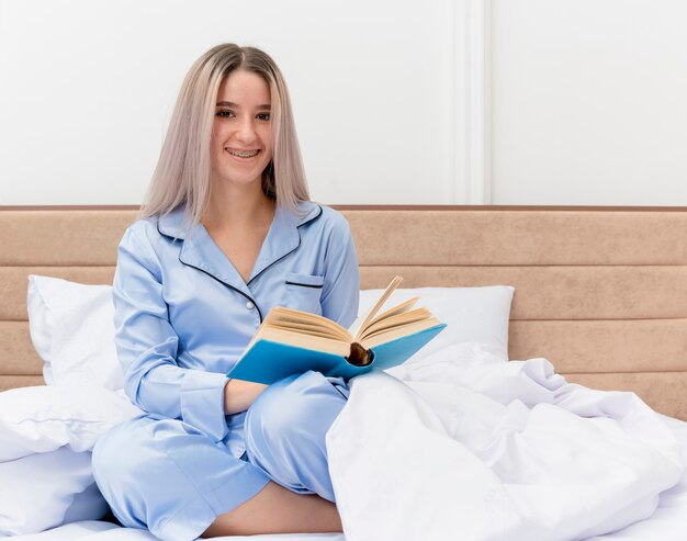 Jovem mulher bonita de pijama azul sentada na cama com um livro sorrindo e uma carinha feliz descansando no interior do quarto