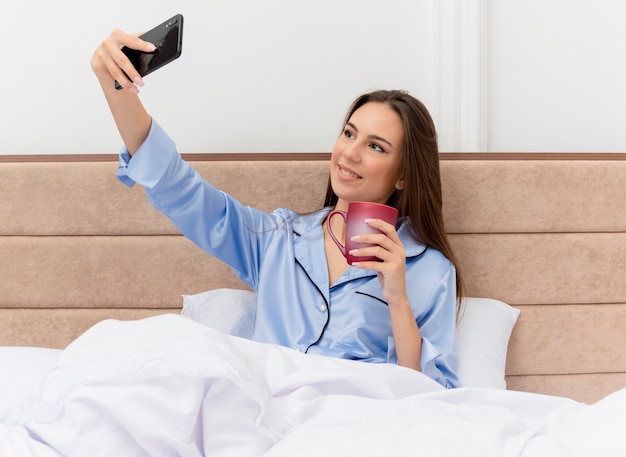 Foto grátis jovem mulher bonita de pijama azul sentada na cama com uma xícara de café fazendo selfie sorrindo com uma cara feliz no interior do quarto na luz de fundo