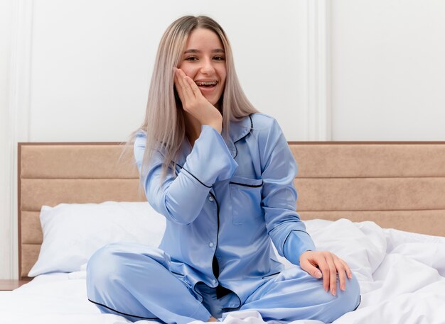 Jovem mulher bonita de pijama azul sentada na cama olhando para a câmera feliz e surpresa no interior do quarto na luz de fundo