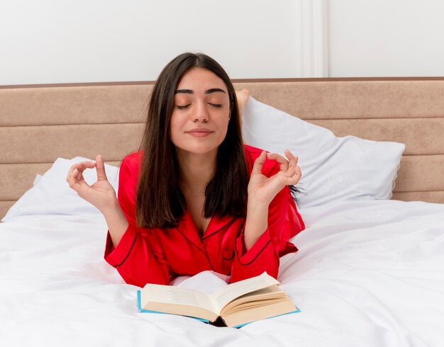 Jovem mulher bonita de pijama vermelho deitada na cama com o livro e os olhos fechados relaxando fazendo gestos de meditação com os dedos no interior do quarto sobre fundo claro