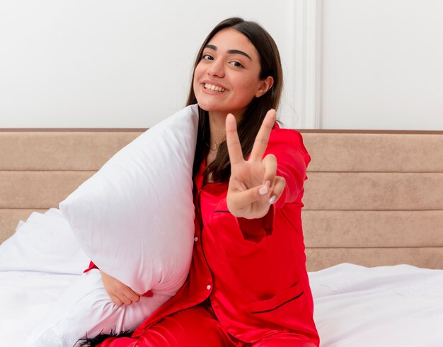 Jovem mulher bonita de pijama vermelho sentada na cama com travesseiro olhando para a câmera, sorrindo, mostrando o sinal-V no interior do quarto com luz de fundo