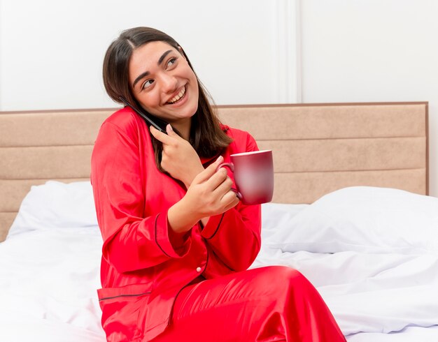 Jovem mulher bonita de pijama vermelho sentada na cama com uma xícara de café falando no celular, sorrindo no interior do quarto sobre fundo claro