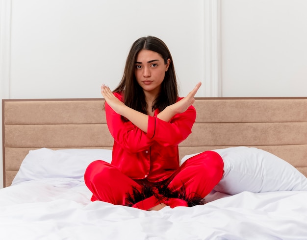 Foto grátis jovem mulher bonita de pijama vermelho sentada na cama fazendo gesto de pare, cruzando as mãos com uma cara séria no interior do quarto