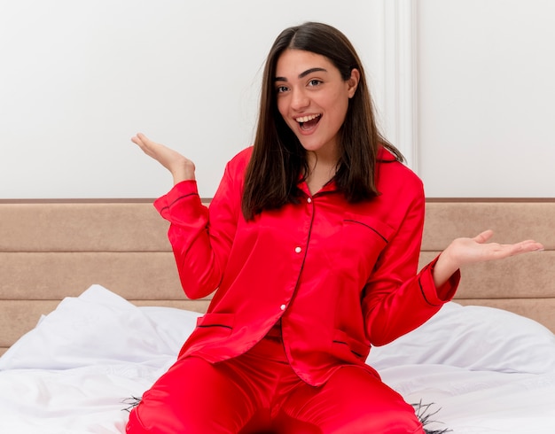 Foto grátis jovem mulher bonita de pijama vermelho sentada na cama feliz e animada, sorrindo alegremente no interior do quarto sobre fundo claro