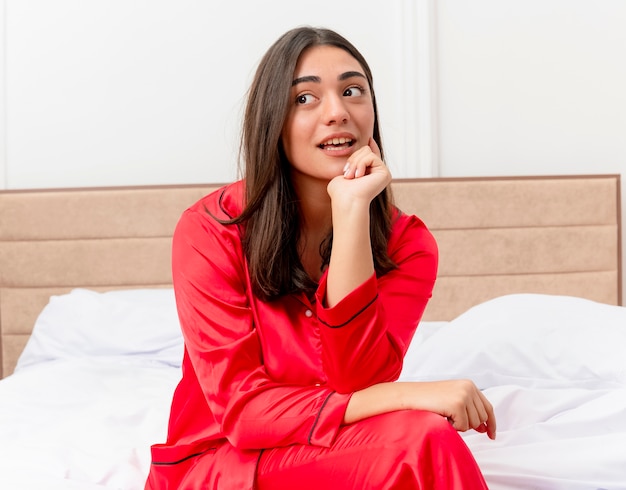 Jovem mulher bonita de pijama vermelho sentada na cama olhando para o lado, pensando positivo, sorrindo no interior do quarto sobre fundo claro