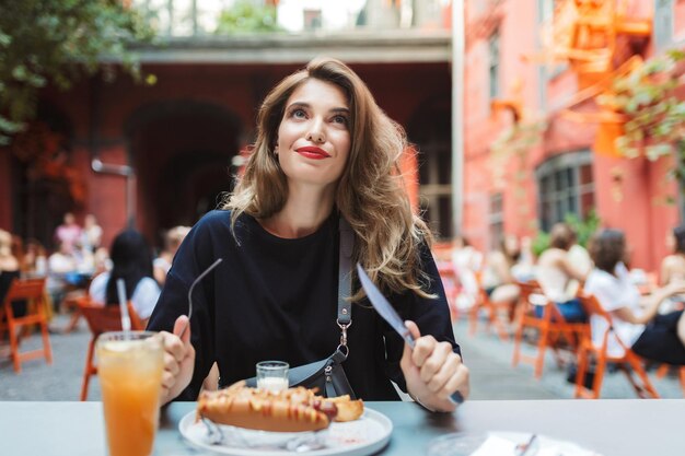 Jovem mulher bonita de vestido preto segurando garfo e faca nas mãos com comida na mesa sonhadoramente olhando para cima enquanto passa o tempo no acolhedor pátio do café