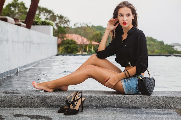 Jovem mulher bonita e sexy sentada na piscina descalça, pernas longas e magras bronzeadas, sapatos de salto alto, camisa preta, shorts jeans, bolsa, relaxante, estilo verão, férias, detalhes de close-up