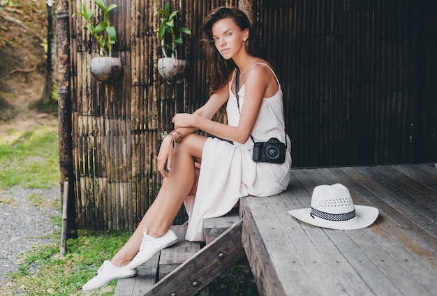 Jovem mulher bonita em férias tropicais na Ásia, estilo verão, vestido boho branco, tênis, câmera fotográfica digital, viajante, chapéu de palha, sorrindo, boho