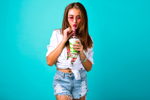 Jovem mulher bonita em miniburta jeans bebendo um smoothie saboroso, roupa vintage, óculos de sol com maquiagem