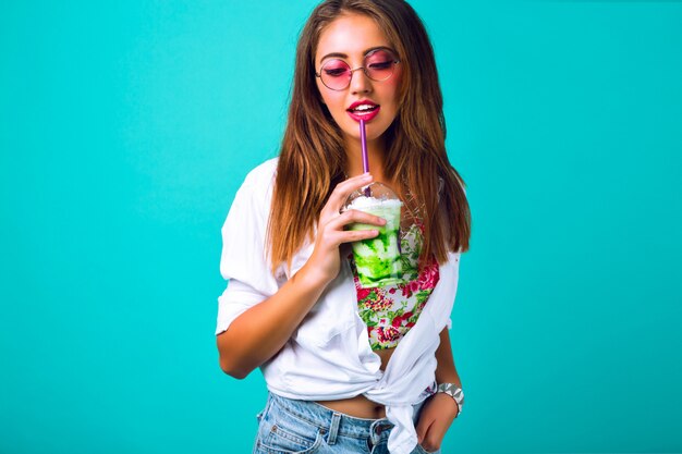 Jovem mulher bonita em miniburta jeans bebendo um smoothie saboroso, roupa vintage, óculos de sol com maquiagem