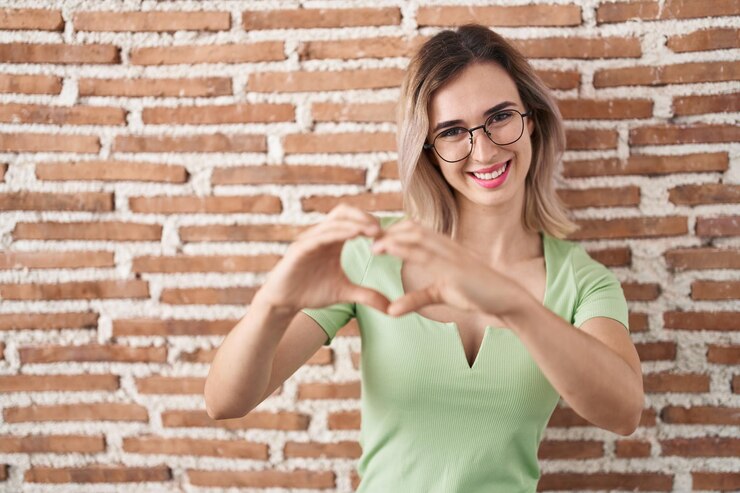 Foto grátis jovem mulher bonita em pé sobre a parede de tijolos sorrindo apaixonadamente fazendo forma de símbolo de coração com as mãos. conceito romântico.