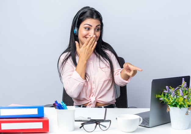 Jovem mulher bonita em roupas casuais com fones de ouvido e microfone sentada à mesa com laptop apontando com o dedo indicador para a tela sorrindo alegremente sobre fundo branco trabalhando no escritório