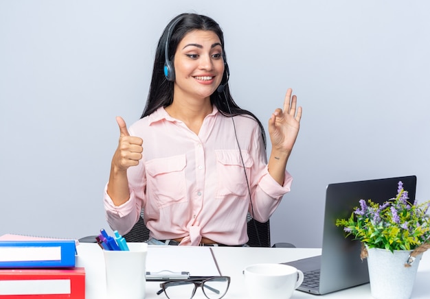 Foto grátis jovem mulher bonita em roupas casuais com fones de ouvido e microfone sentada à mesa com laptop feliz e sorrindo sobre uma parede branca trabalhando no escritório