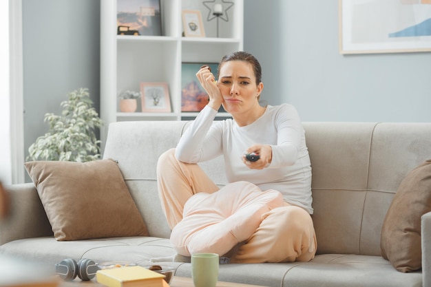 Jovem mulher bonita em roupas casuais, sentado em um sofá no interior de casa comendo biscoito segurando o controle remoto olhando para a câmera confuso e descontente passando o fim de semana em casa