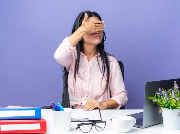 Jovem mulher bonita em roupas casuais usando fone de ouvido com microfone cobrindo os olhos com a mão sentada à mesa com o laptop sobre a parede azul, trabalhando no escritório