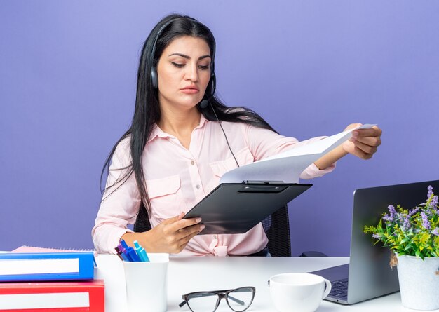 Foto grátis jovem mulher bonita em roupas casuais, usando fone de ouvido com microfone, segurando a prancheta com páginas em branco, parecendo confiante sentada à mesa com o laptop sobre a parede azul, trabalhando no escritório