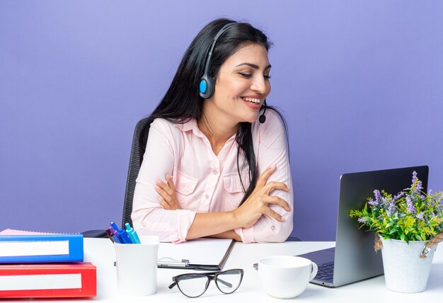 Jovem mulher bonita em roupas casuais usando fone de ouvido com microfone, sentada à mesa com laptop olhando para a tela sorrindo alegremente tendo videochamada em azul