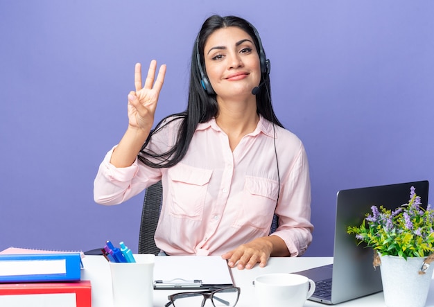 Jovem mulher bonita em roupas casuais usando fone de ouvido com microfone sorrindo confiante mostrando o número três com os dedos sentados à mesa com o laptop sobre a parede azul, trabalhando no escritório