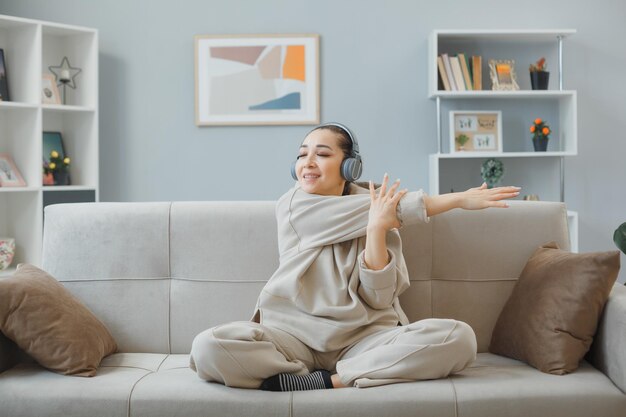 Jovem mulher bonita em roupas de casa sentada em um sofá no interior de casa com fones de ouvido relaxantes felizes e positivos esticando-se fazendo exercícios