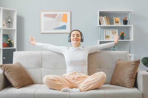 Jovem mulher bonita em roupas para casa, sentado em um sofá no interior de casa com fones de ouvido feliz e positivo relaxante passando o fim de semana em casa