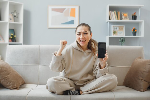jovem mulher bonita em roupas para casa, sentado em um sofá no interior de casa segurando o punho de aperto do smartphone feliz e animado, regozijando-se com seu sucesso