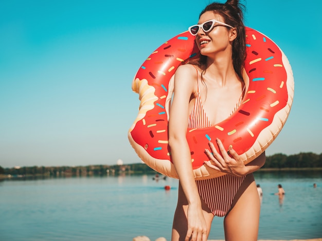 Jovem mulher bonita em trajes de banho e óculos de sol posando na praia com rosquinha inflável