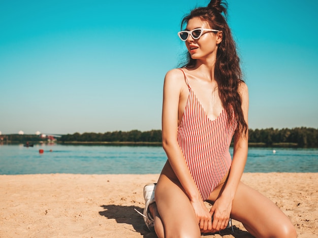 Jovem mulher bonita em trajes de banho e óculos de sol posando na praia