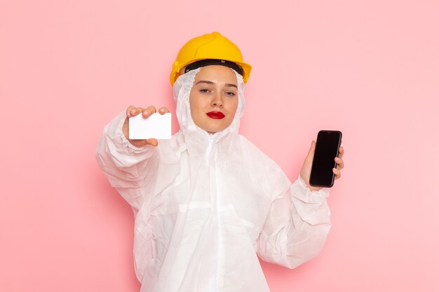 jovem mulher bonita em um terno branco especial e capacete amarelo segurando o telefone e um cartão rosa