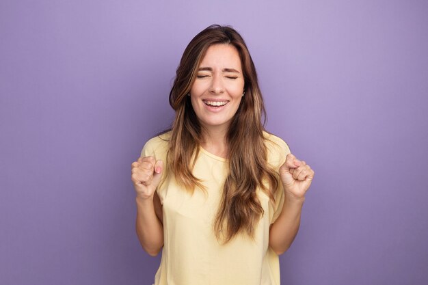 Jovem mulher bonita em uma camiseta bege feliz e animada comemorando seu sucesso cerrando os punhos em pé sobre o roxo
