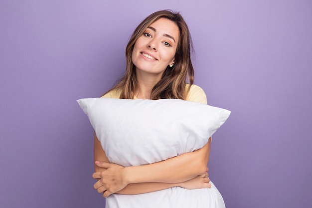 Foto grátis jovem mulher bonita em uma camiseta bege segurando um travesseiro branco, olhando para a câmera com um sorriso no rosto em pé sobre o roxo