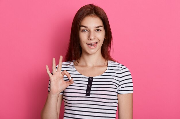 Jovem mulher bonita fazendo sinal de ok com os dedos e mostrando sua língua, com expressão engraçada, posando isolada sobre parede rosa.