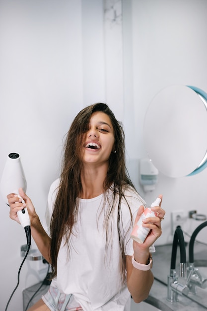 Jovem mulher bonita no banheiro segurando um secador de cabelo e uma pequena garrafa, vista de perto